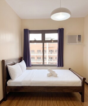 A single bed with white linens and a rolled towel, a window with purple curtains, and an air conditioning unit on the wall. - Premier Double Room with Balcony and Manila Bay View, Pool View or MOA View (Pasay City)