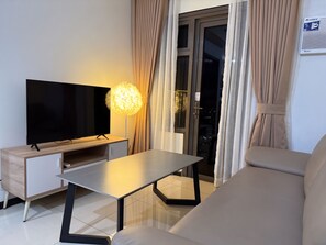A modern living room with a flat-screen TV on a wooden stand, a glass-top coffee table, a sofa, and a view of the city at night through a large window with curtains. - Premier Double Room with Balcony and Manila Bay View, Pool View or MOA View (Pasay City)