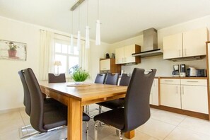 A dining area with a wooden table and black chairs, a window with curtains, and a kitchenette in the background. - Holiday home in Zingst with swimming pool (Zingst)