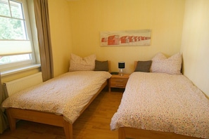 Ein Zimmer mit zwei Einzelbetten, einem Nachttisch mit Lampe, einem Bild an der Wand und einem Fenster mit Vorhängen.
