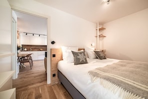 A modern bedroom with a large bed, a bedside table, and a view into a kitchen area. - Houchma – Charming 4-Guest | Mt Blanc View | Spa | Lifts 400m (Les Houches)