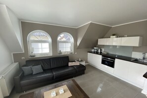 A modern living room with a black leather sofa, a coffee table, and a kitchen area with white cabinets and a black countertop. - Klipper (Captain Parow Holiday Residence) (Zingst)