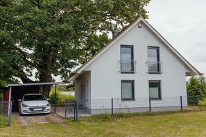 A two-story house with a car parked in the driveway, a fenced yard, and a large tree in the background. - Lakefront property, bathing bay (Krakow am See)