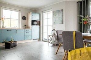 A kitchen with light blue cabinets, a dining area with a grey chair, and a window with a view of birds. - Holiday home with 2 bedrooms (Fuhlendorf)