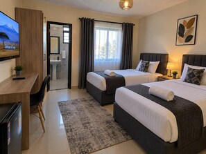 A hotel room with two beds, a desk, a chair, a TV, and a bathroom visible through an open door. - bedsitter r, (Juja)