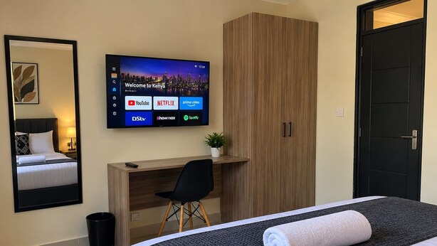 A hotel room with a bed, a desk, a television displaying streaming services, a wardrobe, and a door. - bedsitter r, (Juja)