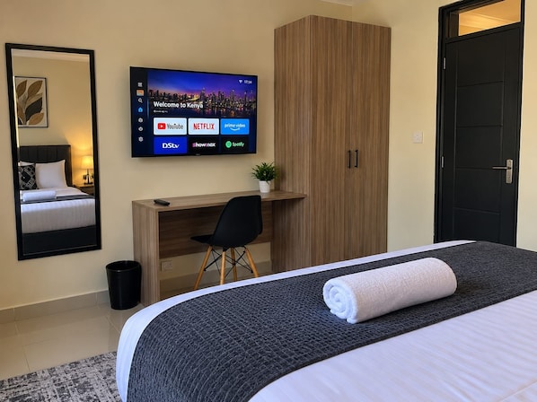 A hotel room with a bed, a desk, a television displaying streaming services, a wardrobe, and a door. - bedsitter r, (Juja)