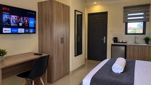 A hotel room with a desk, a bed with a rolled towel, a TV displaying Netflix content, a door, and a kitchenette. - bedsitter r, (Juja)