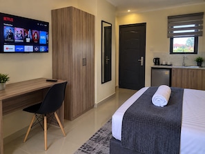 A hotel room with a desk, a bed with a rolled towel, a TV displaying Netflix content, a door, and a kitchenette. - bedsitter r, (Juja)