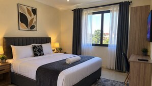 A neatly made bed with a dark headboard, a bedside table with a lamp, a large window with curtains, and a television on a wooden cabinet. - bedsitter r, (Juja)