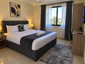 A neatly made bed with a dark headboard, a bedside table with a lamp, a large window with curtains, and a television on a wooden cabinet. - bedsitter r, (Juja)