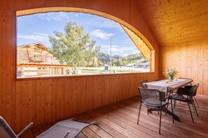 En interiör med träinredning, ett stort, välvt fönster, ett bord med stolar och utsikt över en byggnad och ett träd utanför.