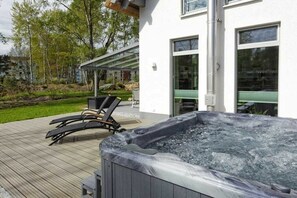 Ein Whirlpool auf einer Holzterrasse mit Liegestühlen, einer überdachten Veranda und einem Gebäude mit großen Fenstern.
