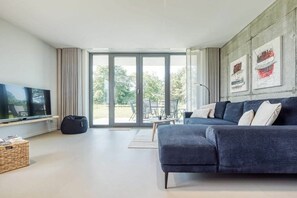 A modern living room with a large flat-screen TV, a dark-colored sofa, and a sliding glass door leading to an outdoor area. - Holiday home with 1 bedroom (Fuhlendorf)