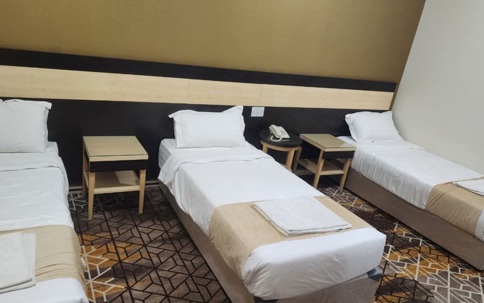 Een hotelkamer met drie eenpersoonsbedden, een klein nachtkastje met een lamp en een telefoon aan de muur.