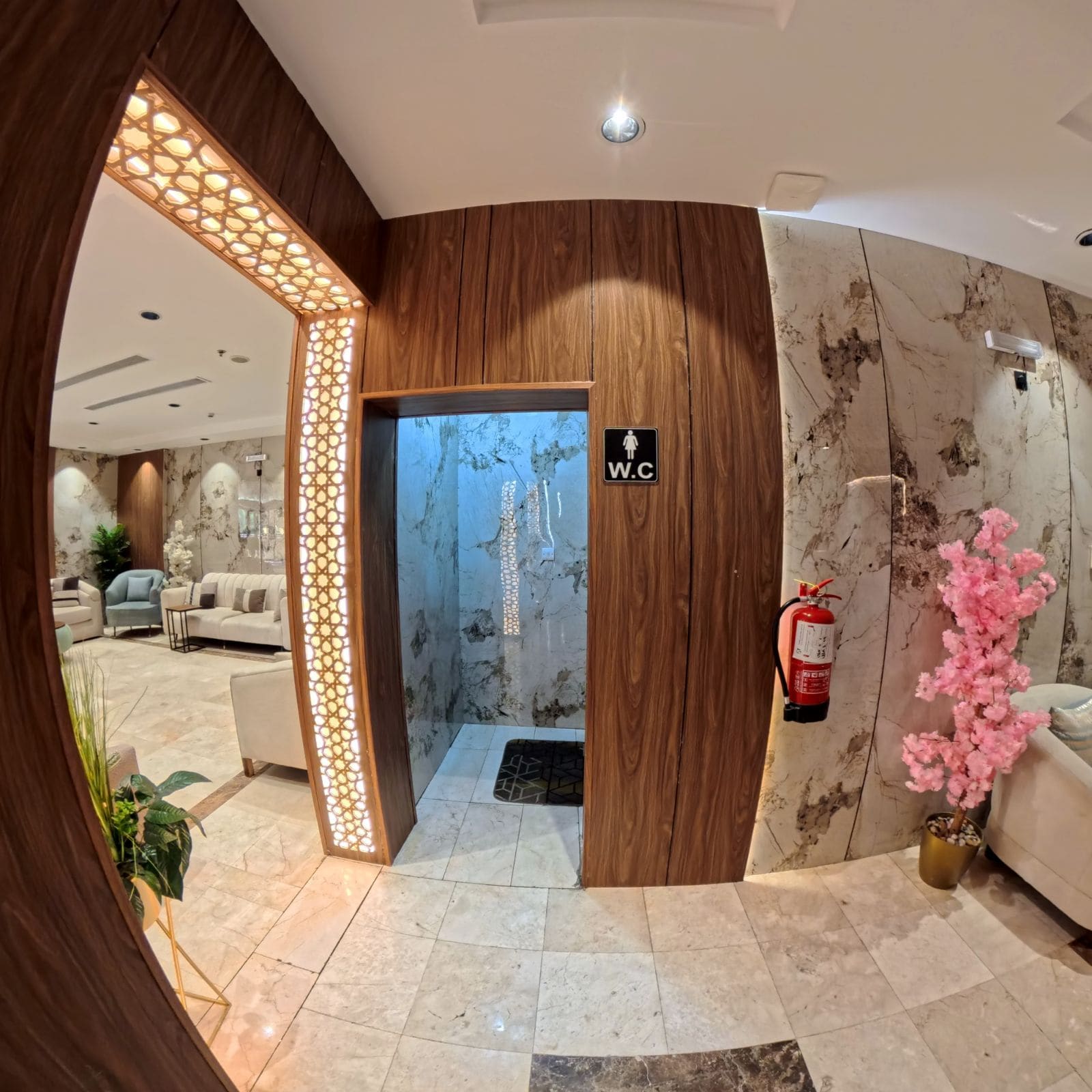 Un vestíbulo de hotel con un mostrador de recepción de madera, un letrero para el baño de mujeres, un extintor y una planta en maceta.