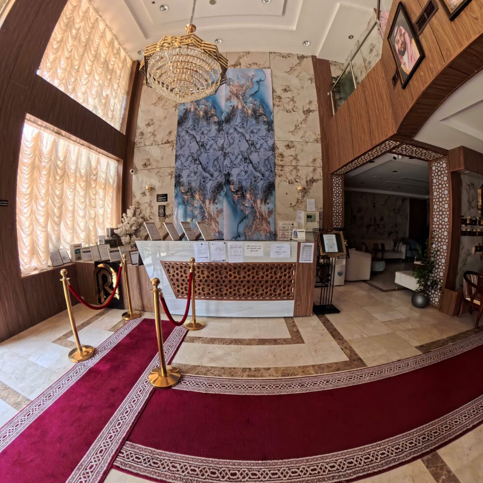 Zona de recepción con pared de mármol, un cuadro abstracto de gran tamaño, una araña y una alfombra roja que conduce a la recepción.