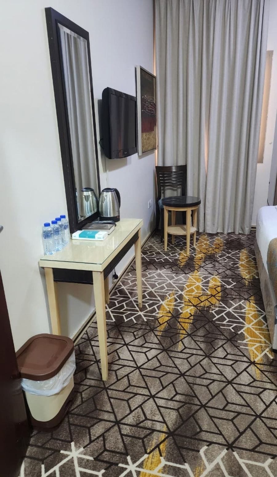 Quarto de hotel com cama, escrivaninha e cadeira, televisão, janela com cortinas e piso estampado.