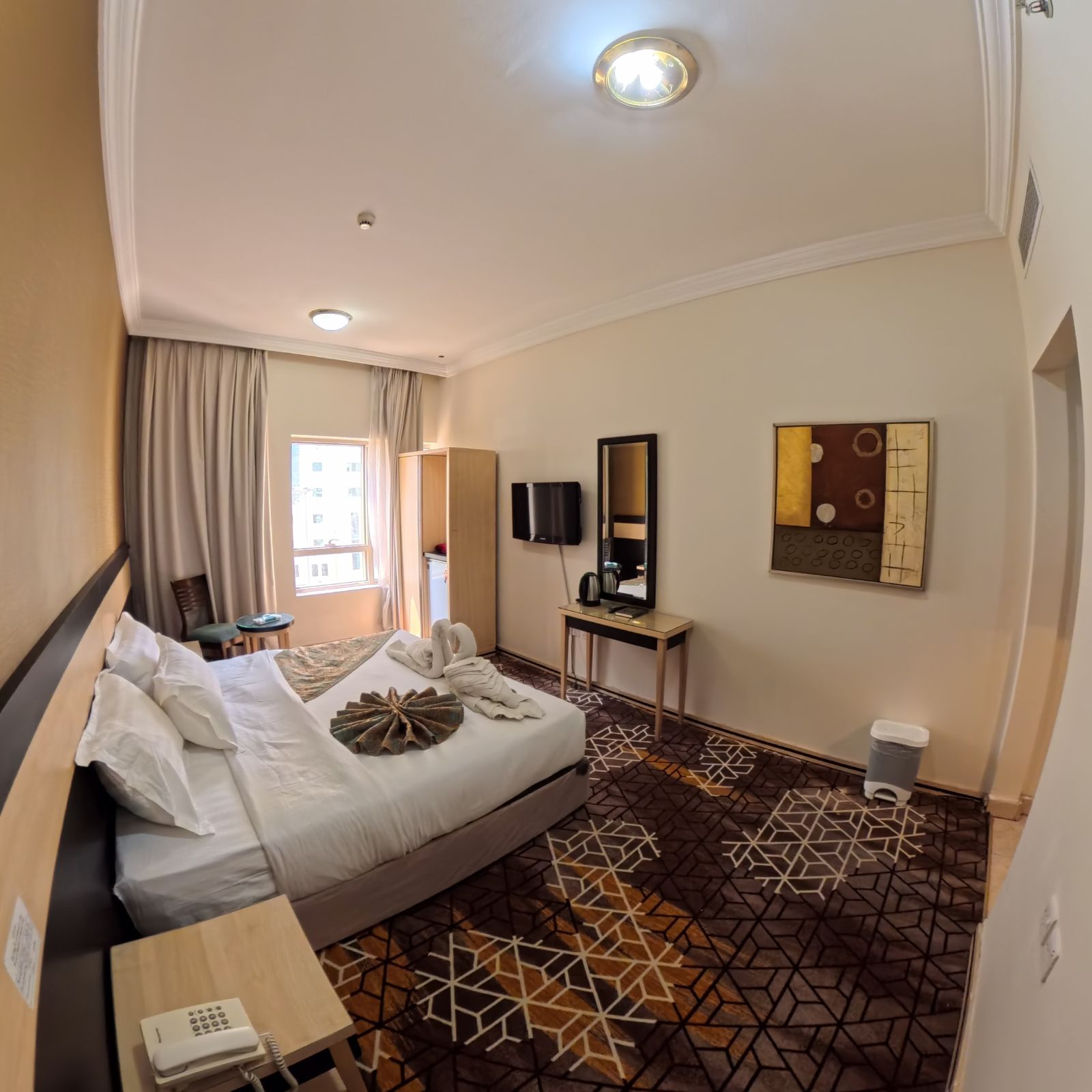Habitación de hotel con cama, mesita de noche, escritorio con teléfono, espejo y televisor montado en la pared.