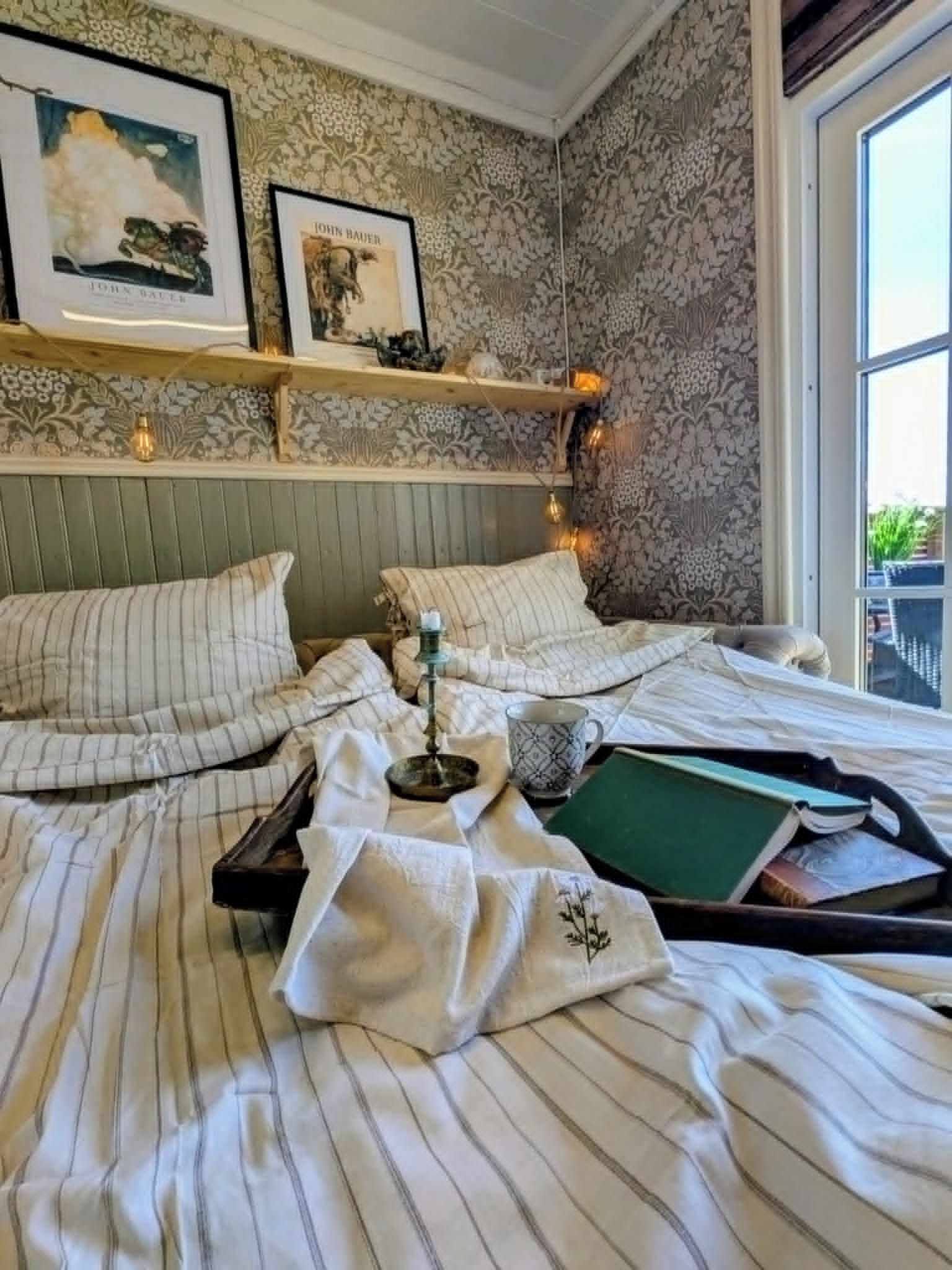 Una cama bien hecha con ropa de cama a rayas, una mesita de noche con una lámpara y una ventana con vistas a la vegetación.