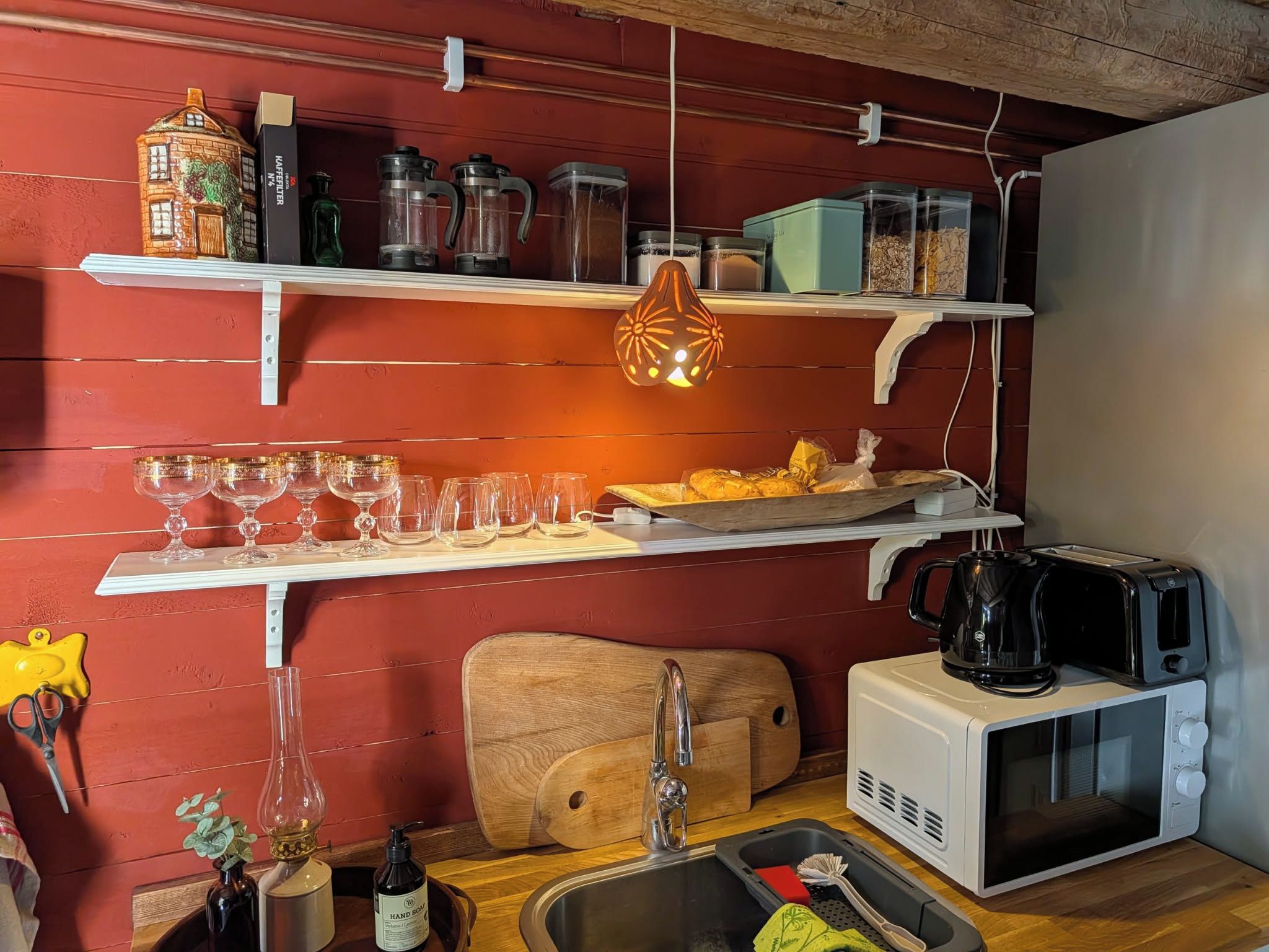 Una cocina con pared roja, repisas blancas, una lámpara colgante, un tostador, un microondas y un lavamanos.