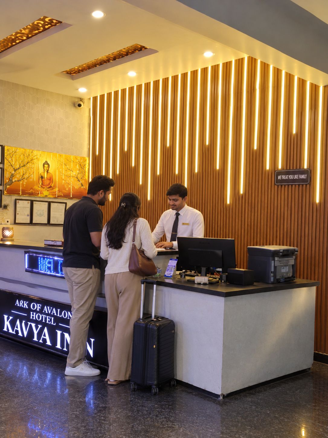 Una recepción con pantalla digital, un cartel que indica 'ARK OF AVALON HOTEL KAVYA INN' y una pared de madera con listones verticales.