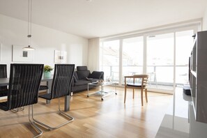 A modern living room with a sofa, chairs, a dining table, and a large window. - Apartment 9, Südstrasse 1 (Norderney)
