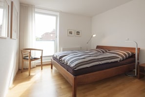 A bedroom with a wooden bed, a chair, a bedside table, and a window with curtains. - Apartment 9, Südstrasse 1 (Norderney)