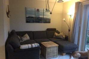 Ein Wohnzimmer mit einem dunkelgrauen Sofa, einem kleinen Hocker, einem Holztisch und einem Gemälde eines Leuchtturms an der Wand.