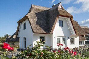 A house with a unique triangular roof design, white exterior walls, and a garden with various flowers. - Holiday home with 2 bedrooms (Fuhlendorf)