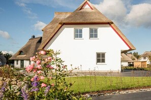 A white house with a thatched roof and a red trim, surrounded by a well-maintained lawn and flowering plants. - Holiday home with 2 bedrooms (Fuhlendorf)