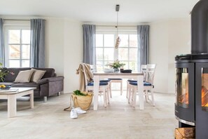 A modern living room with a sofa, dining table, chairs, a fireplace, and large windows with curtains. - Holiday home with 2 bedrooms (Fuhlendorf)