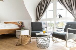 Ein modernes Schlafzimmer mit einem Bett, zwei Sesseln, einem kleinen Tisch und einem Fenster mit Vorhängen.