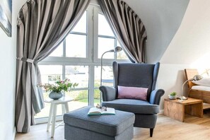 Une chambre moderne avec une grande fenêtre, un fauteuil gris, une petite table avec une plante et une table de chevet avec une lampe.