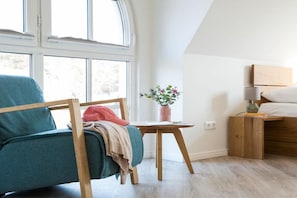 Ein Zimmer mit einem blaugrünen Sessel, einem Nachttisch aus Holz mit einer Lampe, einem kleinen Topfpflanzen und einem Bett mit Holz Kopfteil.