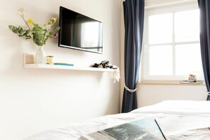 A hotel room with a bed, a mounted TV, a window with curtains, a shelf with a vase of flowers, and a small table with a candle. - Holiday home with 2 bedrooms (Fuhlendorf)