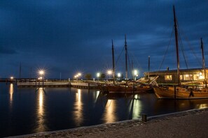 A marina at night with boats docked, a wooden building with lights, and a calm water surface reflecting the lights. - Holiday home with 2 bedrooms (Fuhlendorf)