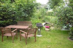 Une table et des chaises en bois, disposées dans un jardin agrémenté d’un bac à fleurs en métal et d’un élément décoratif aquatique.