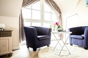 A room with two dark blue armchairs, a white table with a vase of flowers, a lamp, and a window with curtains. - ||--------------------Reed Warbler 30------------------|| (Fuhlendorf)