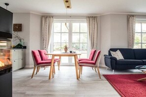 A living room with a fireplace, a dining table with chairs, a sofa, and a large window with curtains. - Holiday home with 2 bedrooms (Fuhlendorf)