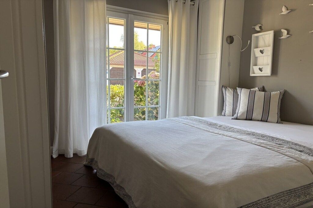 A bedroom with a large bed, white curtains, and a view of a house through a glass door.