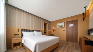 A hotel room with a large bed, a wooden headboard, a bedside table, a desk with a chair, a door, and a wall-mounted shelf with a picture. - Shili Huating Hongpu Hotel (Guangzhou)