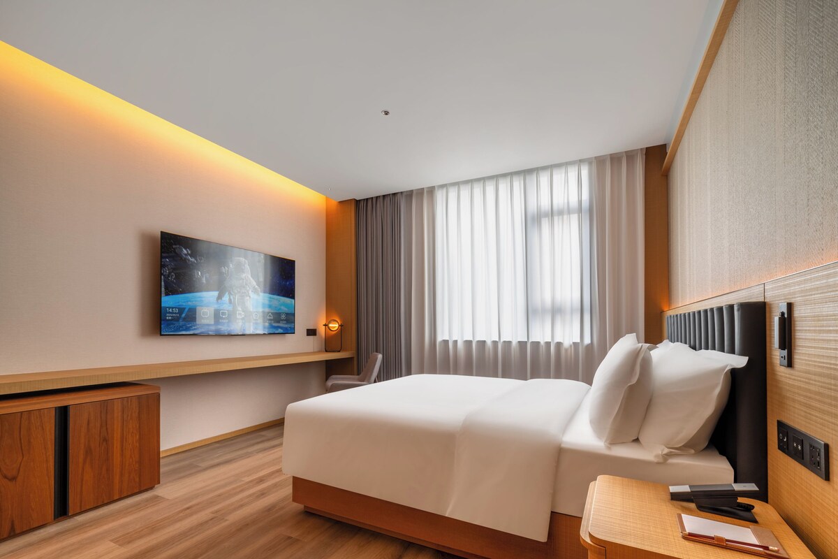 A hotel room with a bed, a desk, a chair, a television mounted on the wall, and a window with curtains.