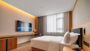 Una camera d'albergo con un letto, una scrivania, una sedia, una televisione a parete e una finestra con tende.