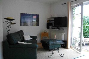 Ein Wohnzimmer mit einer Couch, einem Couchtisch, einem Fernseher und einem Fenster mit Blick auf Grünflächen.