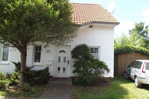 Ein weißes Haus mit Ziegeldach, einer Satellitenschüssel, einem Auto auf der Auffahrt und einem Baum davor.