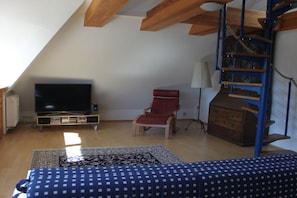 Ein Zimmer mit Fernseher, einem roten Sessel, einem blauen Sofa, einer Holzkiste und einer Wendeltreppe.