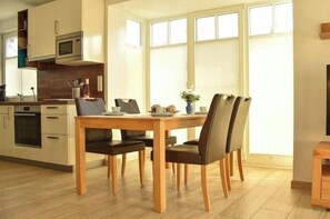 Ein Essbereich mit Holztisch und -stühlen, im Hintergrund eine Küche und große Fenster.