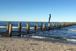 Ein Strand mit Holzpfählen, die ins Wasser ragen, und einer Statue einer Person auf einem der Pfähle.
