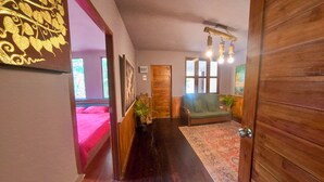 Un pasillo con puerta de madera, un sofá verde, una alfombra estampada y una cama con ropa de cama rosa.